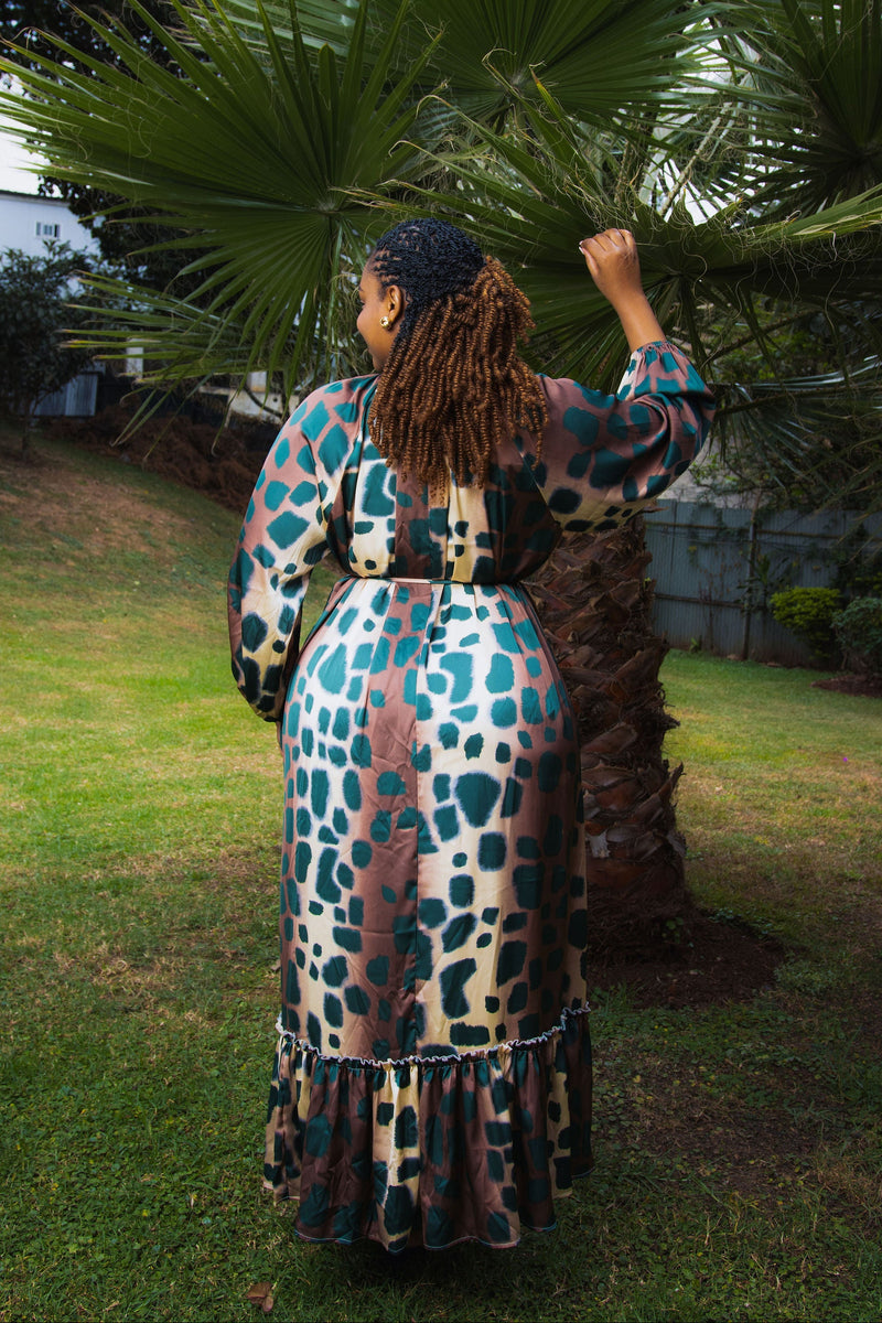 A woman stands outdoors in a grassy yard under a palm tree, wearing a long-sleeve maxi dress with a blue, brown, and beige animal print pattern, viewed from behind.
