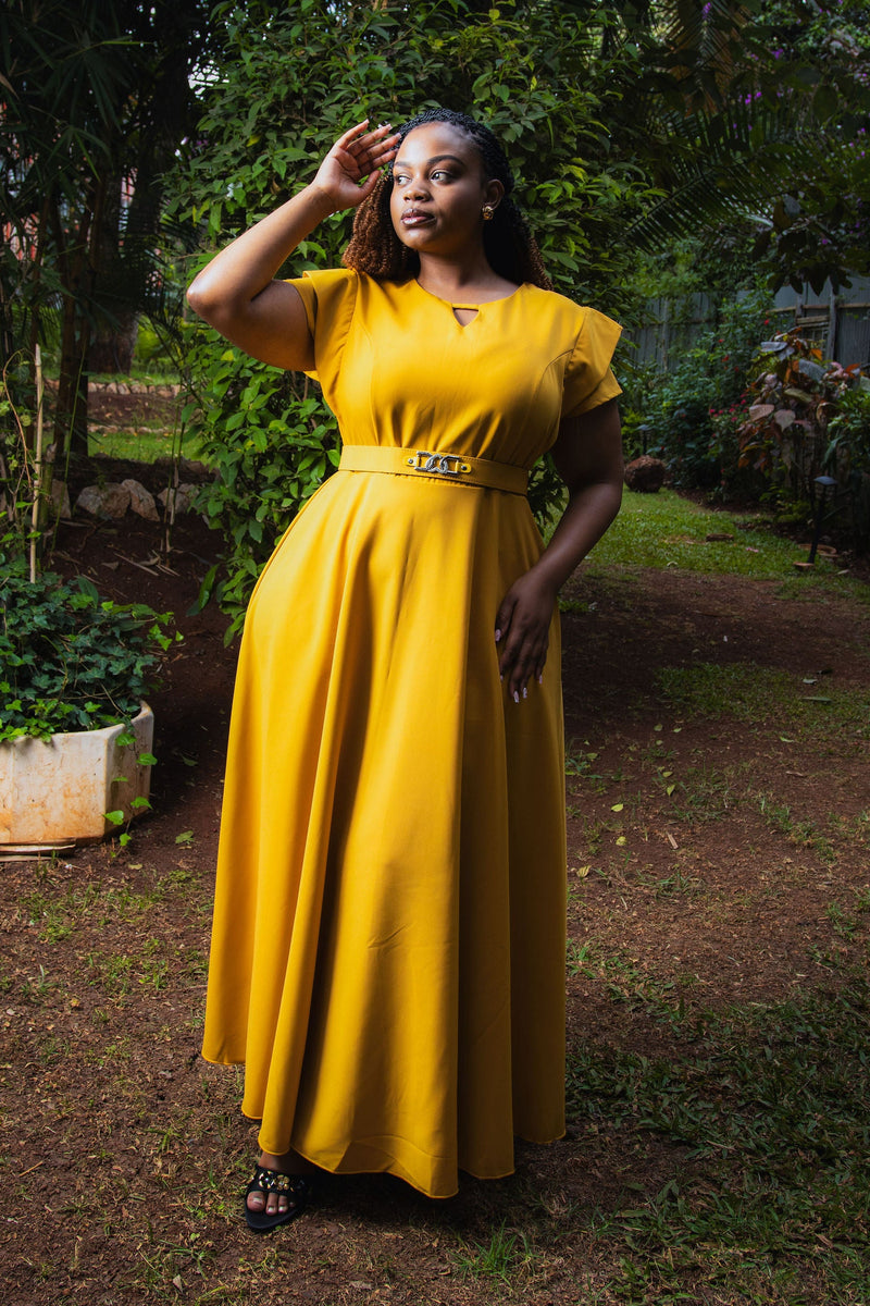 A woman wearing a bright yellow maxi dress with short flutter sleeves and a matching belt stands outdoors in a garden setting.