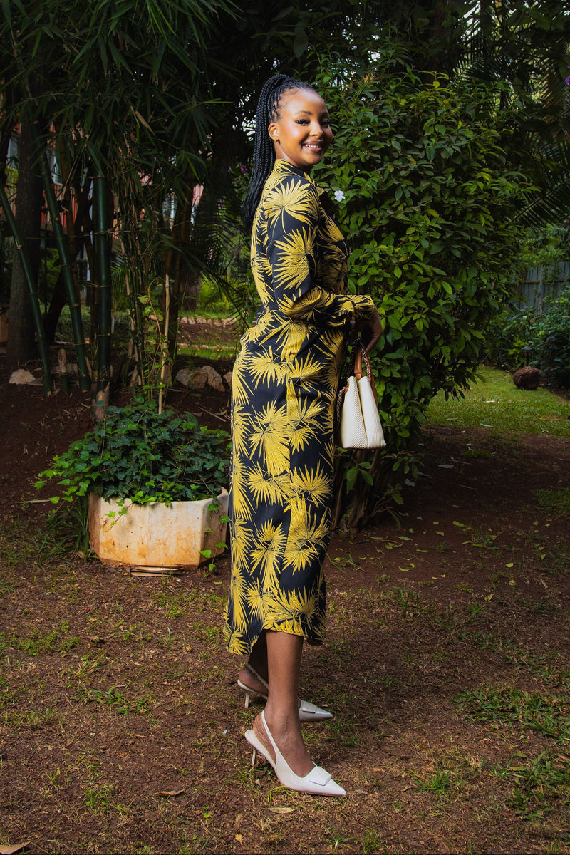 A woman stands outdoors in a garden wearing a long-sleeve midi dress with a bold yellow and black palm leaf pattern, paired with white pointed-toe heels and holding a small white handbag.