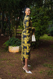 A woman stands outdoors in a garden wearing a long-sleeve midi dress with a bold yellow and black palm leaf pattern, paired with white pointed-toe heels and holding a small white handbag.