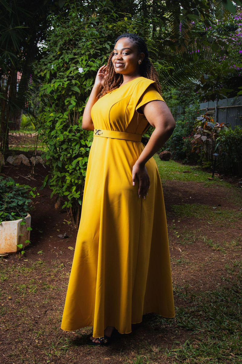 A woman wearing a bright yellow floor-length dress with short sleeves and a matching belt stands outdoors in a garden setting with lush green foliage and a dirt path.