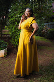 A woman wearing a bright yellow floor-length dress with short sleeves and a matching belt stands outdoors in a garden setting with lush green foliage and a dirt path.